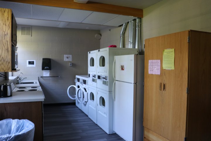 A shared laundry and kitchen space featuring stacked washers and dryers, a refrigerator, and a stove. Cabinets and a microwave are visible on one side of the room, while a bulletin board with notices hangs on the opposite wall.